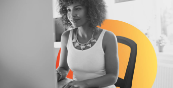 Mulher negra com cabelo crespo sentada em frente ao computador, trabalhando em ambiente de escritório, com fundo em tons de laranja que destacam a imagem em preto e branco.