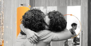 Duas mulheres de cabelos cacheados se abraçam em um ambiente interno, em imagem em preto e branco com elementos gráficos em laranja nas bordas.