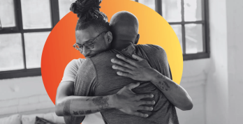 Duas pessoas negras se abraçam de forma acolhedora em um ambiente interno com janelas ao fundo. A imagem está em preto e branco, com um círculo em tons de laranja e amarelo ao fundo destacando a cena e transmitindo sensação de apoio, cuidado e conexão.